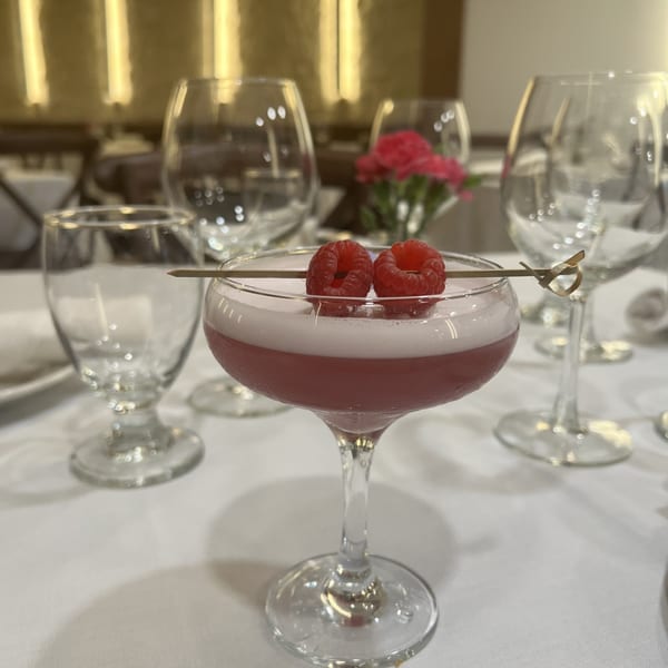 A pink cocktail with foam and two raspberries on a skewer, on a white tablecloth with glasses.