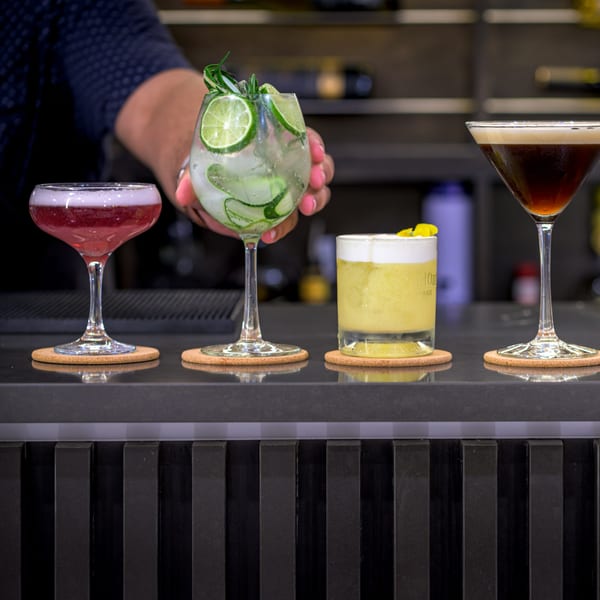 Bartender's hand placing a cucumber and lime cocktail next to three other drinks on a bar.