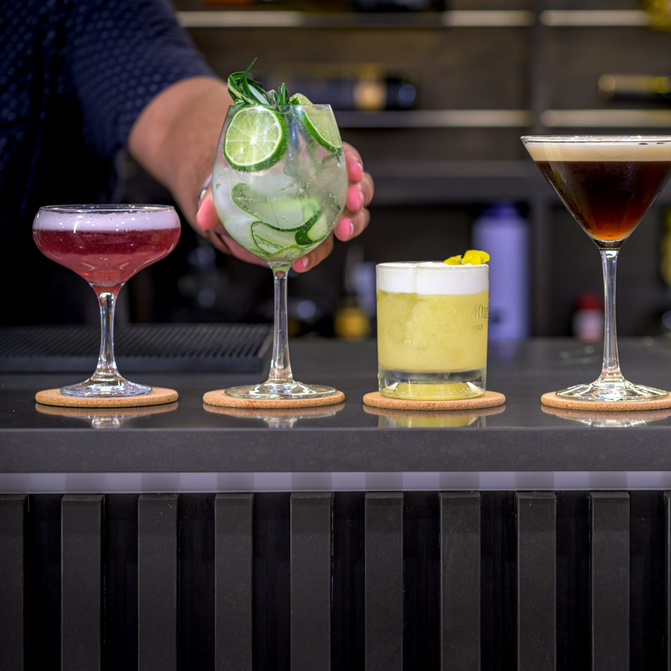 Bartender's hand placing a cucumber and lime cocktail next to three other drinks on a bar.