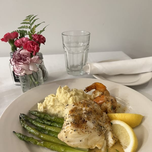 A plate of baked fish with asparagus, mashed potatoes, shrimp, and lemon wedges on a white tablecloth.