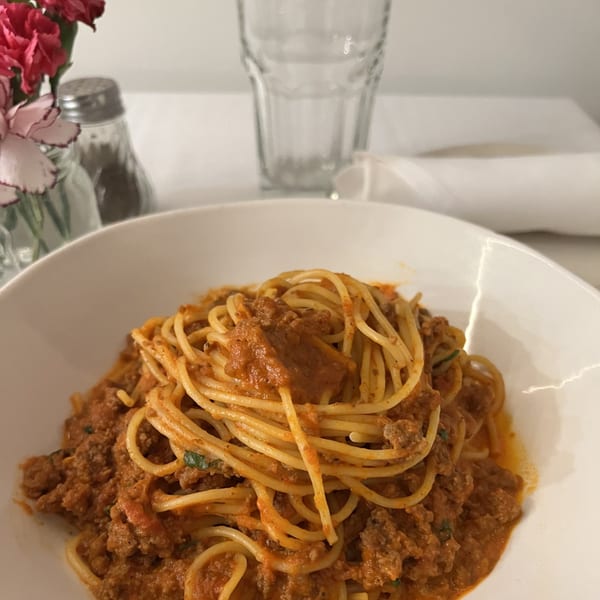 A white bowl filled with spaghetti and meat sauce, with flowers and a glass in the background.