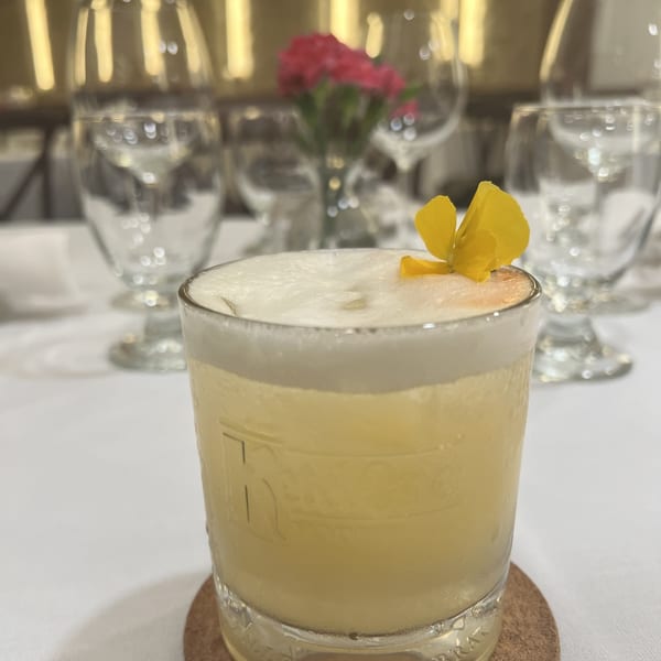 A foamy yellow cocktail garnished with a yellow flower sits on a white tablecloth.