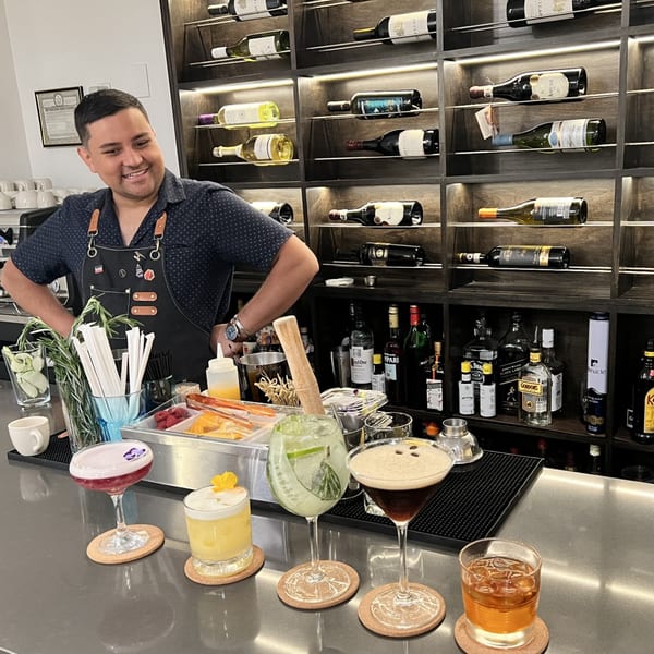 A smiling bartender stands behind a bar with five cocktails and a wine rack in the background.